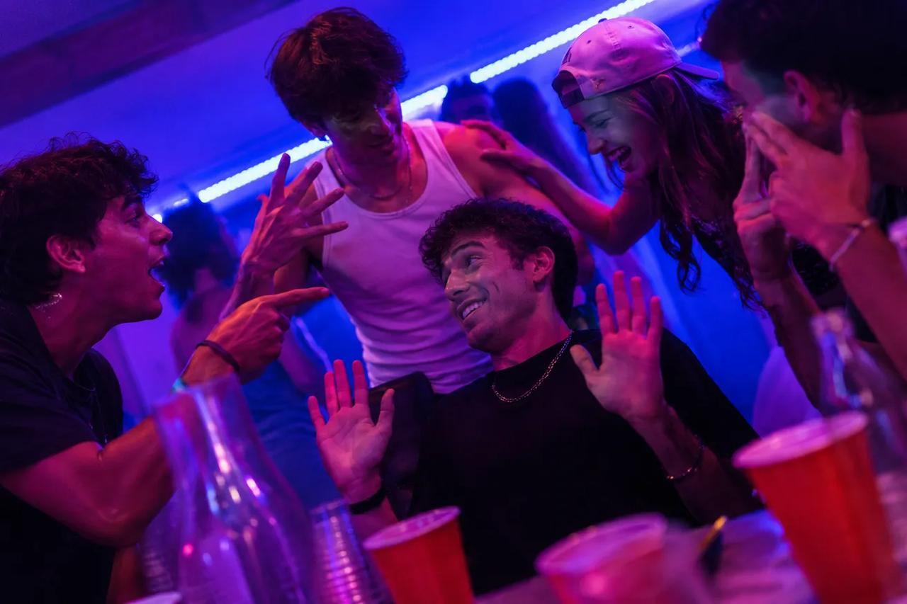 Grupo de amigos jogando Cadeira Quente em uma festa em casa, um amigo no meio rindo enquanto os outros disparam perguntas