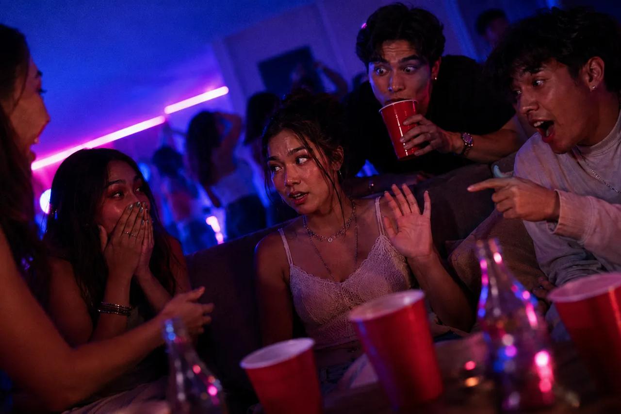 Amigos reunidos ao redor de uma pessoa na cadeira quente, chocados com a resposta dela para uma pergunta polêmica sobre um crush secreto