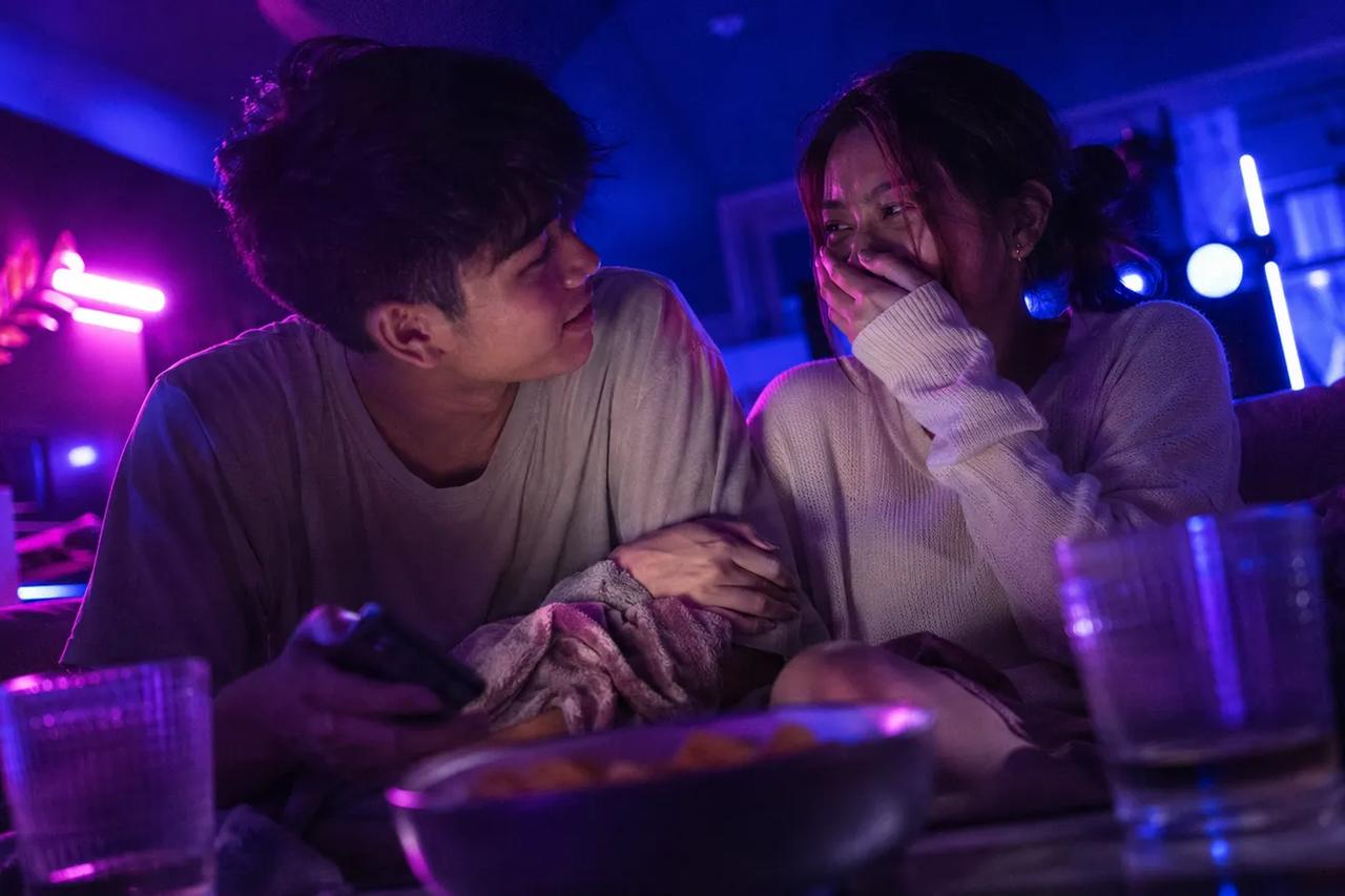 Isang couple na nakaupo sa couch, tumatawa at nag-e-enjoy sa isang masayang quiz game habang nag-da-date night
