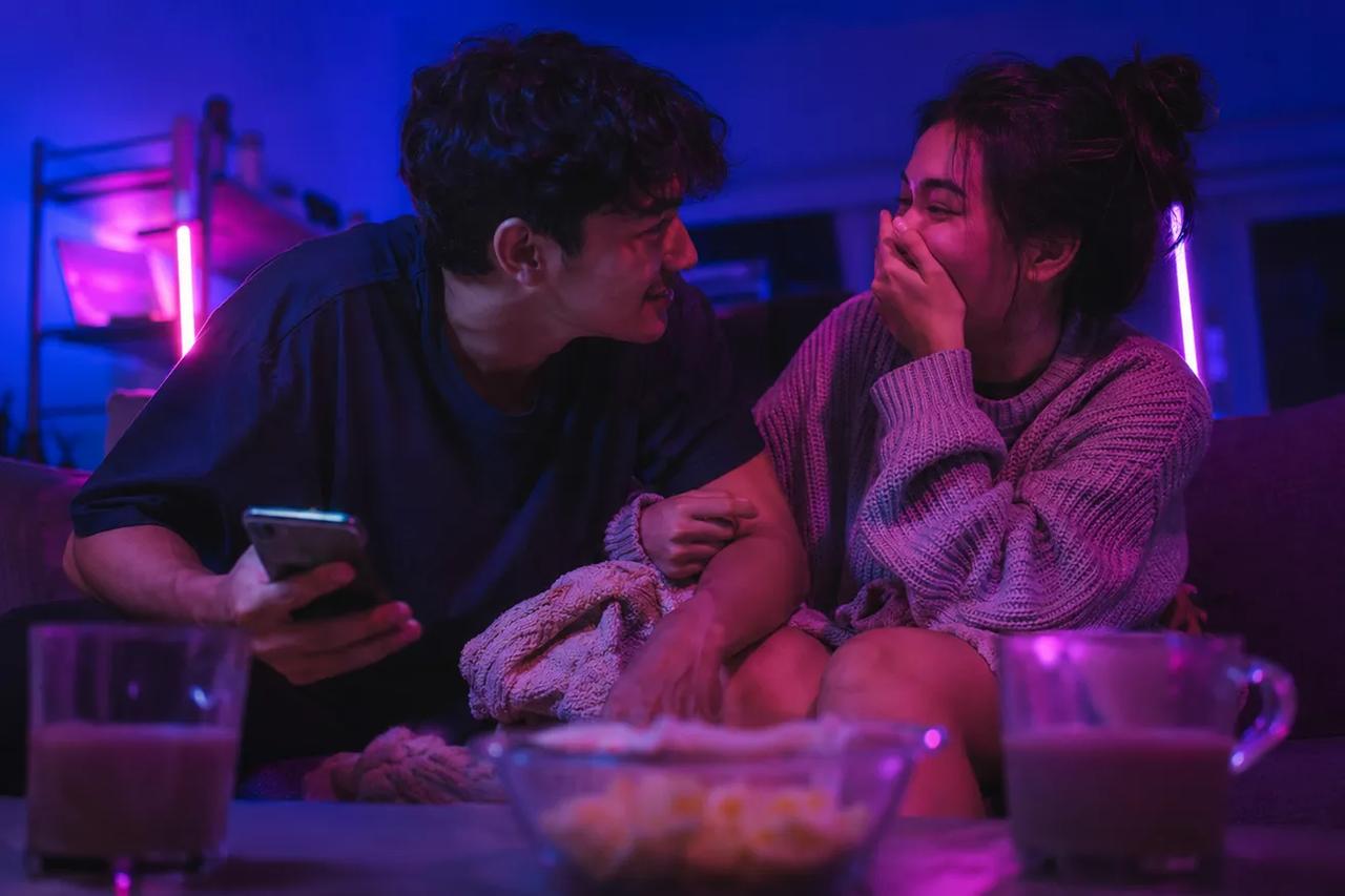 Sepasang kekasih duduk bareng di sofa, ketawa dan nikmatin game kuis seru pas lagi date night