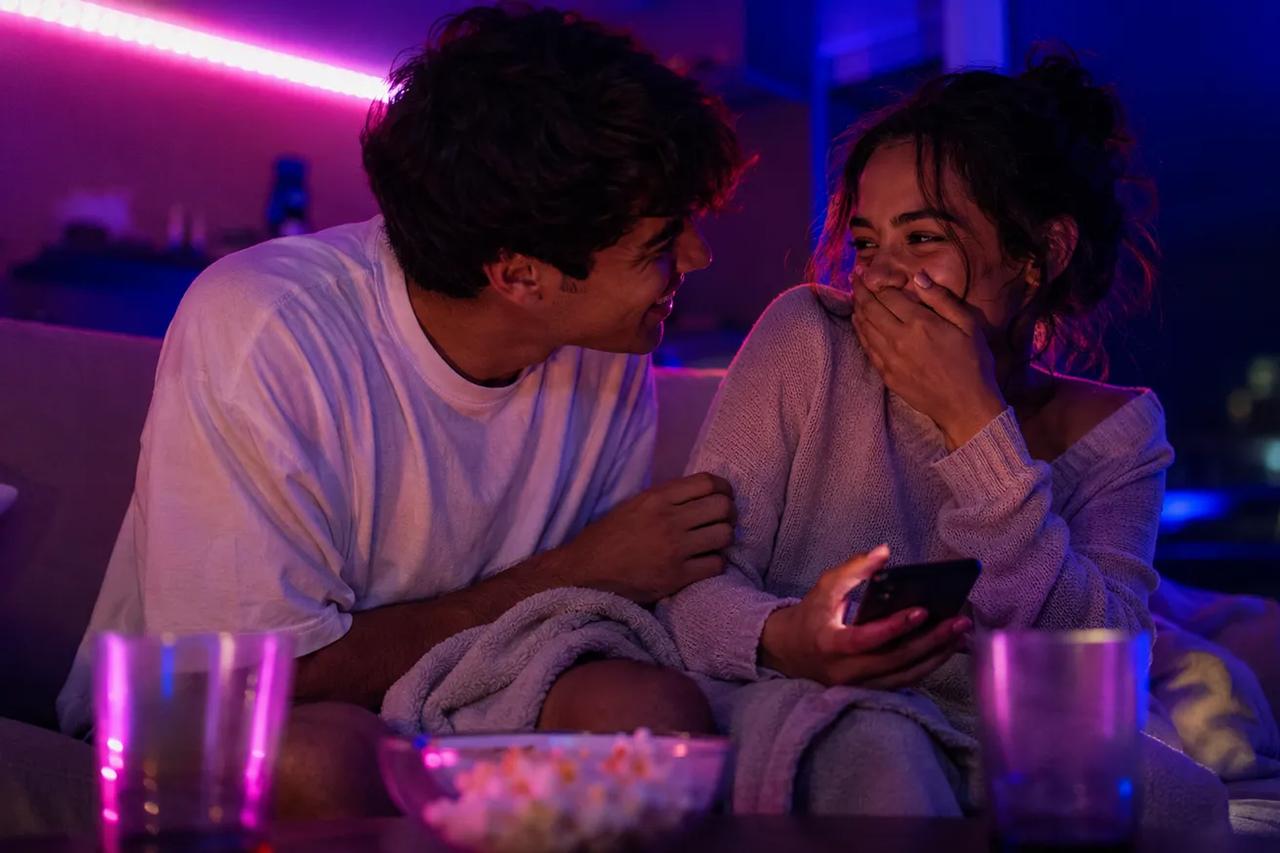 Um casal sentado junto no sofá, rindo e se divertindo com um quiz em uma noite de encontro